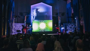 Musicians perform on stage at Firth Court in front of spectacular projected visuals representing particles