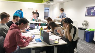 Scene in a classroom with about 9 students working around tables 