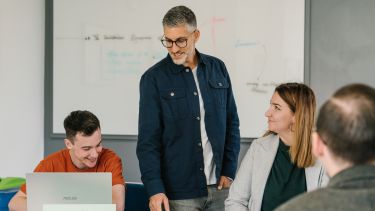 A politics lecturer standing next to a group of students sat at a table in a seminar, speaking to the group and smiling