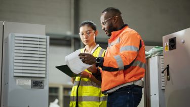 Two people stand in front of engineering equipment, looking at documents in their hands.