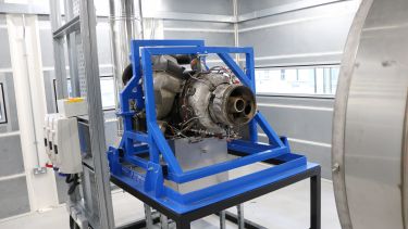 a piece of research equipment - which looks like a jet engine displayed in a blue metal frame - sits in front of a tube in a white room 