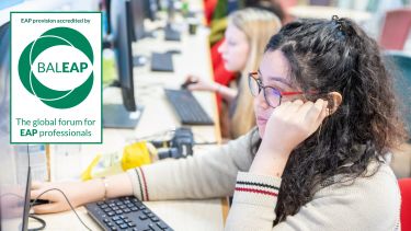 An image showing a female student sat at a desktop computer in a room with other students using computers in the background. The image also contains the BALEAP logo to demonstrate the Pre-sessional English Course is accredited by the BALEAP association of Lecturers in English for Academic Purposes