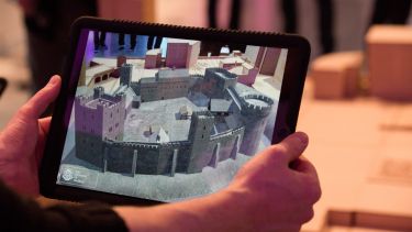 An iPad displaying an augmented reality rendering of Sheffield Castle is held in front of a wooden scale model of the Castlegate site