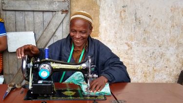 A tailor from rural tanzania sits in front of an old-style sewing machine which has been retrofitted with a new motor. He is smiling and wearing a traditional hat. A smiling local woman sits on a nearby wall over his shoulder.