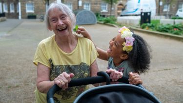 An older woman with grey hair pushing a pram laughs with her grandchild 