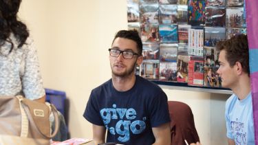 A person sits behind a table, wearing a dark blue t-shirt that says "Give it a Go" in light blue lettering.