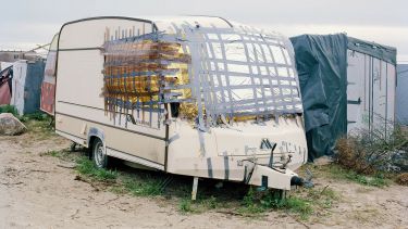 Frontal image of a caravan in a muddy field, The broken windows are covered with blankets stuck on with ducktape 