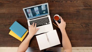 A person sat a table on an online conference call. They have a notebook in front of them with a mug of tea in their hand.