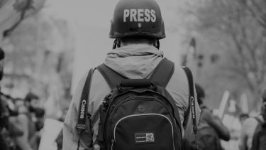 A person with a large rucksack, helmet labelled 'press' and two cameras strapped to their back facing away from the camera. 