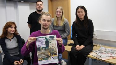 A group of five people sat in a classroom. The man at the front holds up a poster that reads 'blow the whistle on domestic violence'