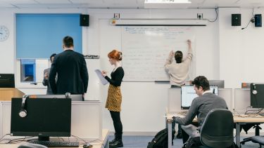A group of students in a classroom - two speaking to a teacher, one writing on a whiteboard, one at a PC.