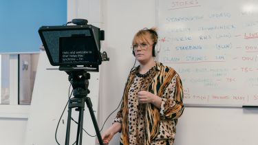 A woman wearing headphones stands next to a teleprompter.