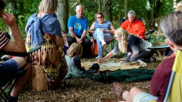 Group of people of mixed ages in a circle, sat in a forest, interacting with "out of the box"