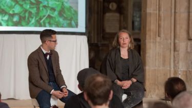 Two individuals sat in front of an audience, discussing slides as shown on a projector screen behind them