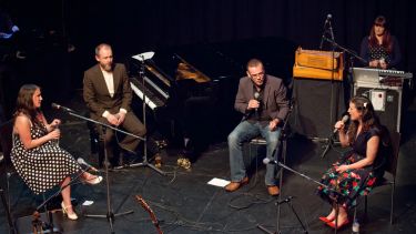 A group of individuals sat on a stage with microphones, ready to sing; one of them is sat at a piano