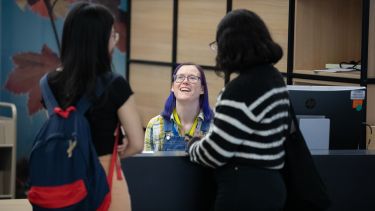 Two people standing at talking to the third person sitting at the Library Welcome Desk