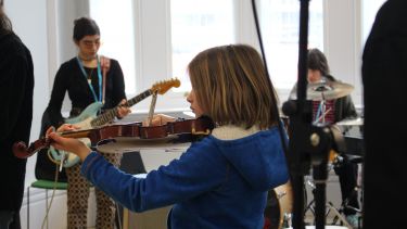 A girl playing the violin in the foreground. Two girls playing the bass guitar and drums in the background.