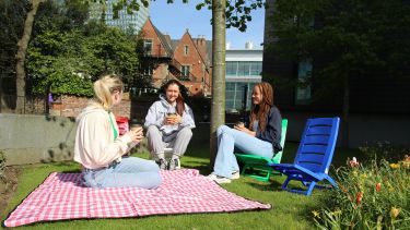Three students sit on deckchairs and a picnic blanket outside the Information Commons