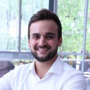 A photo of a man smiling at the camera. He has dark hair and a beard, and is wearing a white collared shirt.