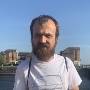 A man with brown hair and a beard. He is wearing a white shirt and standing next to a bridge