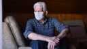 Picture of a man in a checked shirt sat on the edge of a sofa. The man is wearing a paper mask.