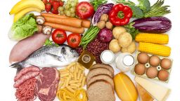 Top view of a white table filled with a large group of different types of food like carbohydrates, protein and dietary fiber. Food included in the composition are dairy products, sausages, minced meat, poultry, fish, bread, pasta, rice, beans, nuts, olive oil, fruits and vegetables