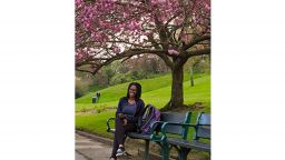 A photograph of Ifedayo Ayodeji sitting on a park bench underneath a tree with pink blossom. 