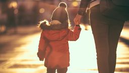 A single parent holds the hand of their young child as they walk down the road.