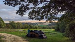 Green farmland and fields stretch out in the distance. In the foreground a man is covering some hay bales with black plastic 