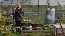 A woman is stood in an allotment next to a raised bed. There is a greenhouse in the background and several tubs of plants growing