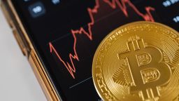 A coin with a Bitcoin symbol lying on a mobile phone screen with an image of stock markets
