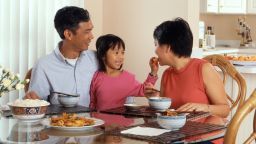 An Asian family, an adult male and female are seated around a table eating a meal with a young female standing in between the adults