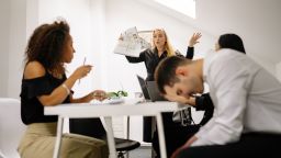 Conflict in the workplace. A man has his head on the table. Two women are seated and talking. One woman is stood up with papers in her hand and arguing.