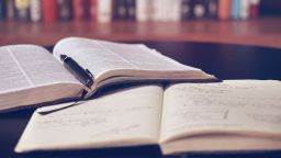 An open printed book, a pen and an open notebook on a table. In the background is a bookshelf.