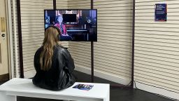 An individual sat on a white bench watching Objectified on a TV screen.
