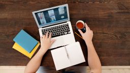 A person sat a table on an online conference call. They have a notebook in front of them with a mug of tea in their hand.