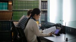 Two people sitting at the desk, reading. Behind them there are shelves filled with books.