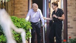 One person helping an elderly person using a walking stick to walk out of a door and down some steps