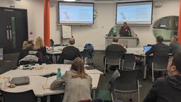 People sat at tables, watching a speaker present slides