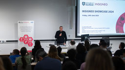 A man wearing a black jumper standing in front of lecturn holding a microphone. He is facing an audience in a lecture theatre.