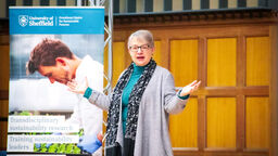 A woman with short grey hair wearing dark framed glasses and a flowing grey cardigan stood with arms outstreched speaking in front of a banner stand in a room with wood panelled walls.